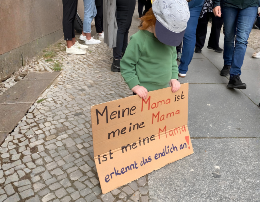 Zwei Mütter sitzen mit ihrem Kleinkind auf einer Bank in einem Gebäude und warten.